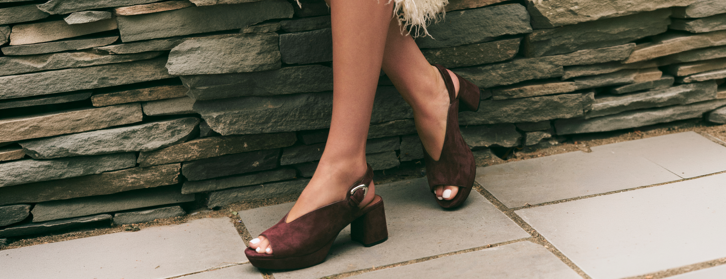 A person wearing dark burgundy peep-toe block-heel pumps with silver buckle straps, standing on a stone patio in front of a stacked stone wall.