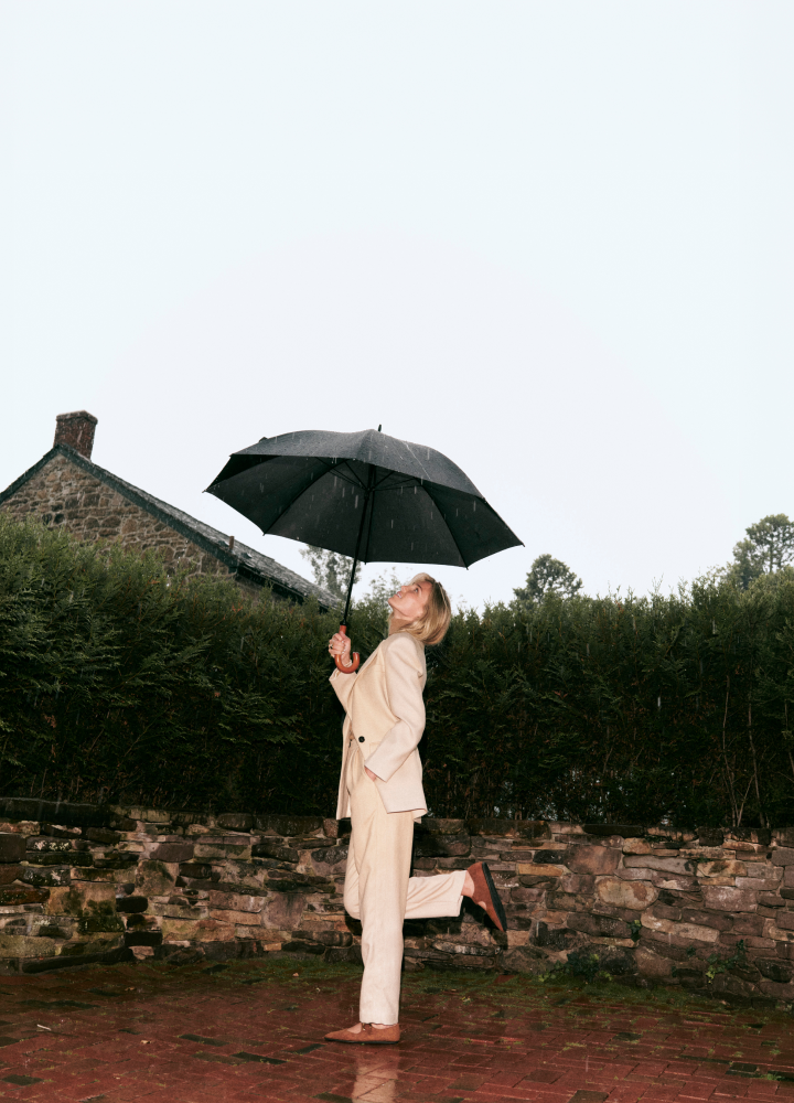 image of woman wearing white trench and pants and tan flats under umbrella in the rain. brick patio with stone and trees in the background. 
