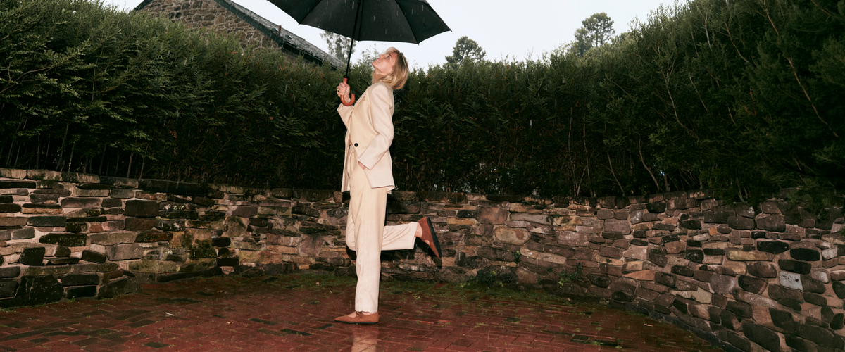 image of woman wearing white trench and pants and tan flats under umbrella in the rain. brick patio with stone and trees in the background. 