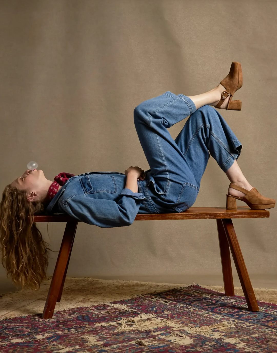 woman laying on a bench blowing bubble gum. wearing a denim outfit with tan suede slingback platform mules