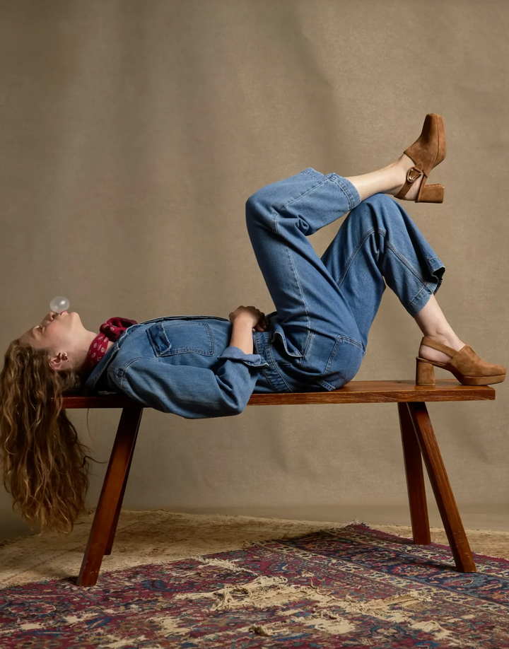 woman laying on a bench blowing bubble gum. wearing a denim outfit with tan suede slingback platform mules