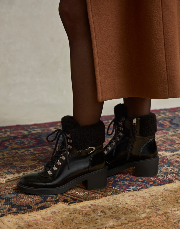 A person wearing black lace-up ankle boots with black faux-fur trim and block heels, standing on a patterned rug. The lower legs are visible, wearing dark tights and a brown skirt.