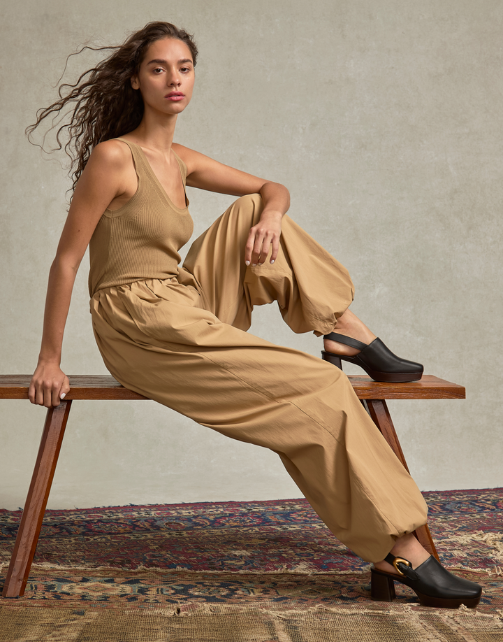 woman sitting on a bench in a studio with vintage carpet wearing tan outfit and black platform slingback mules