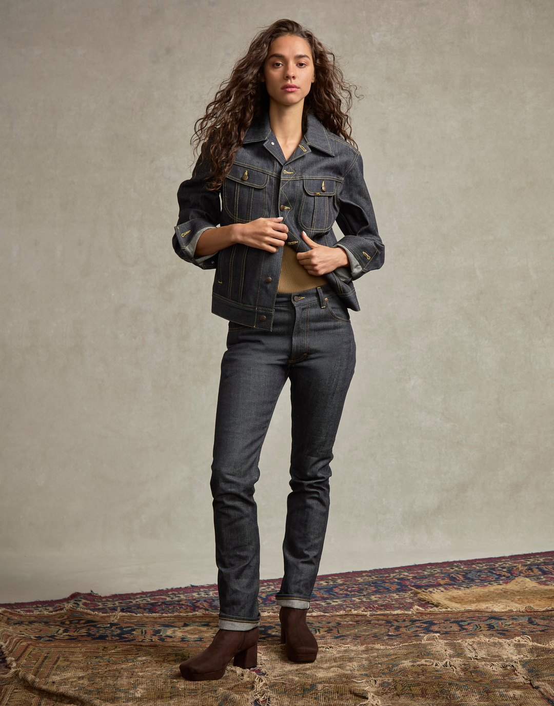 A woman stands in a studio wearing a dark blue denim jacket and matching dark blue jeans, paired with brown ankle boots. She is posed on a patterned rug against a textured beige wall.
