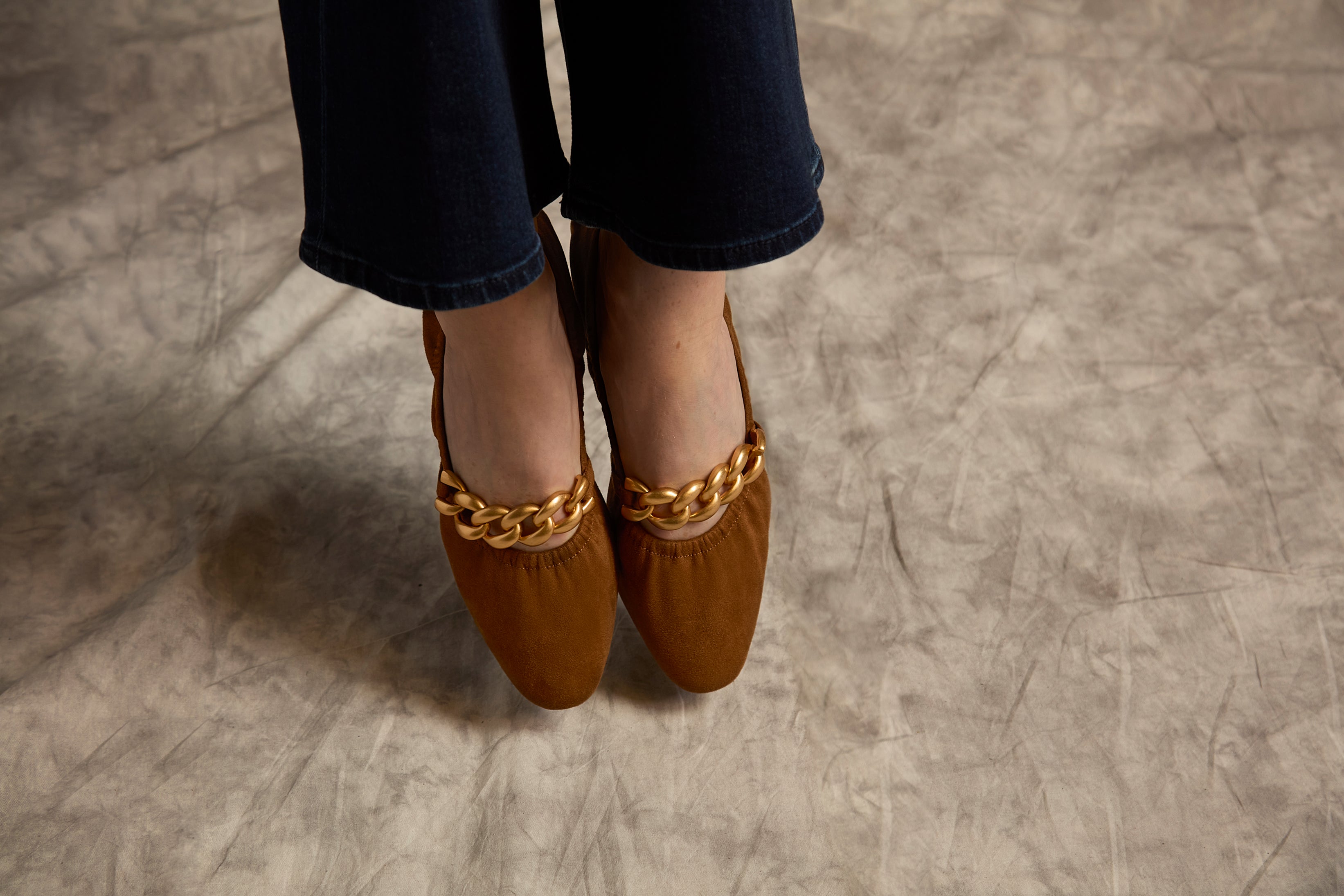 Brown shoes with gold chain details worn by a person on a gray floor.