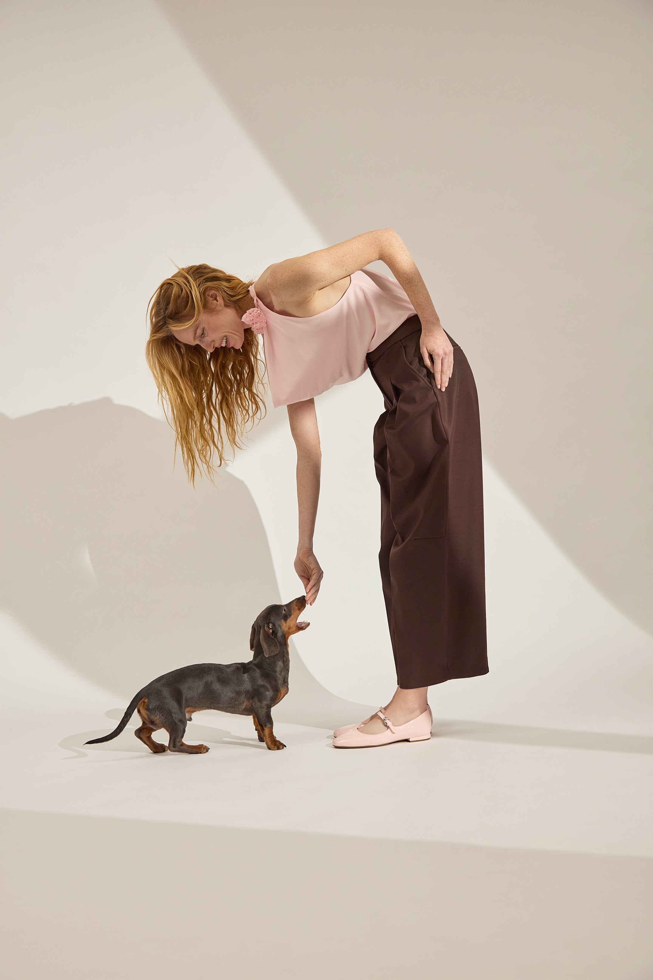 Woman in a pink top and brown pants interacting with a small dog on a white background