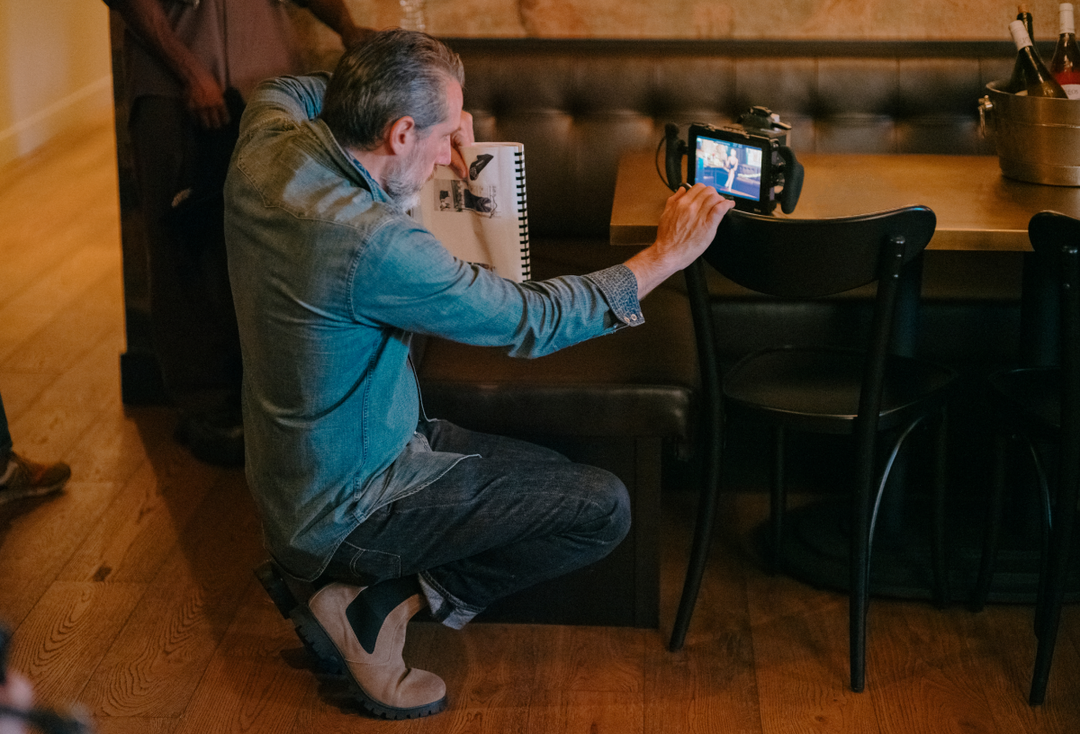 A man in a blue denim shirt and jeans crouches beside a table in a restaurant or cafe, holding a small camera pointed at a menu or notepad.