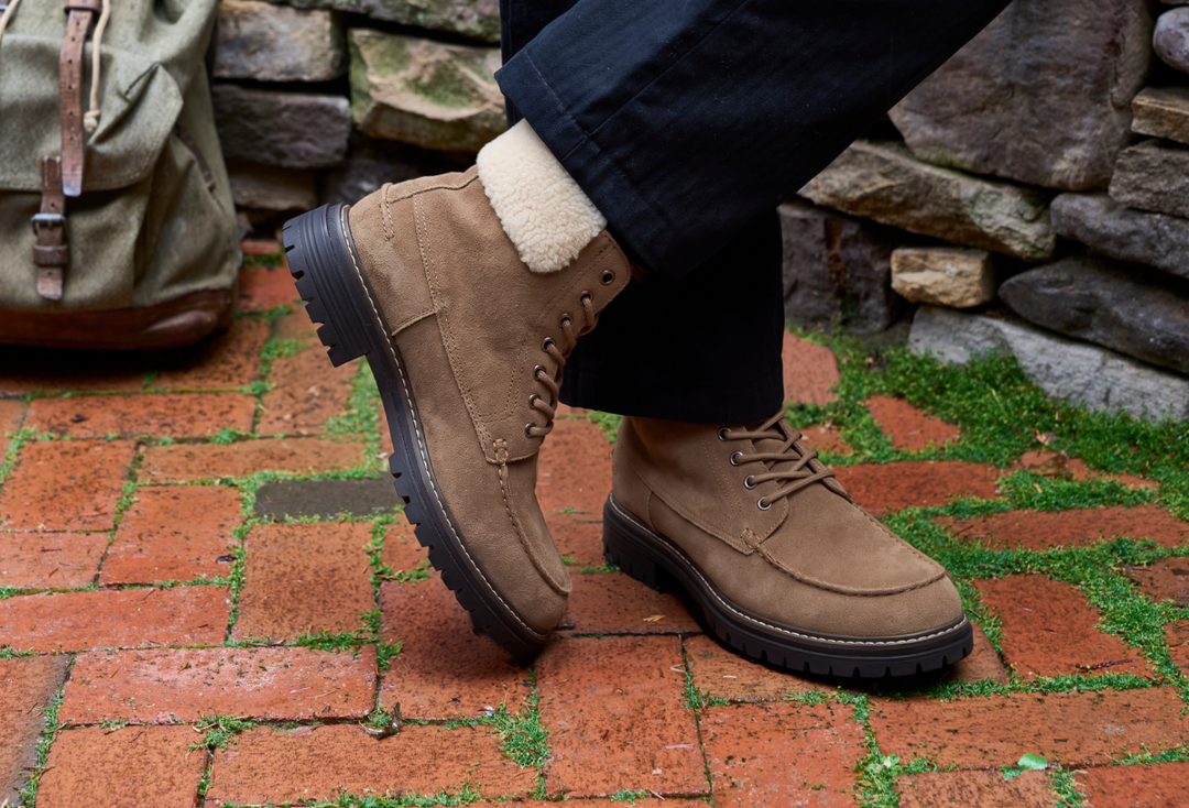 A person wearing tan suede lace-up boots with a cream sherpa lining, standing on a red brick patio with a stone wall and a green backpack in the background.